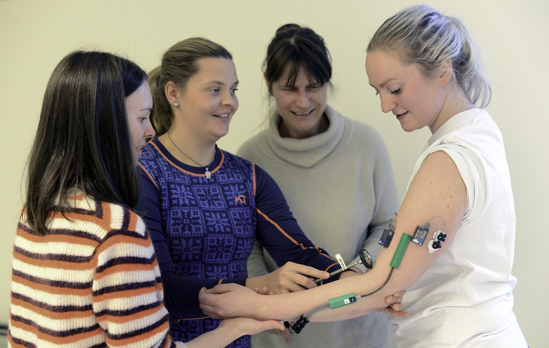 student bruker reflekshammer på medstudent med påmonterte sensorer på arm. Underviser og medstudent ser på.