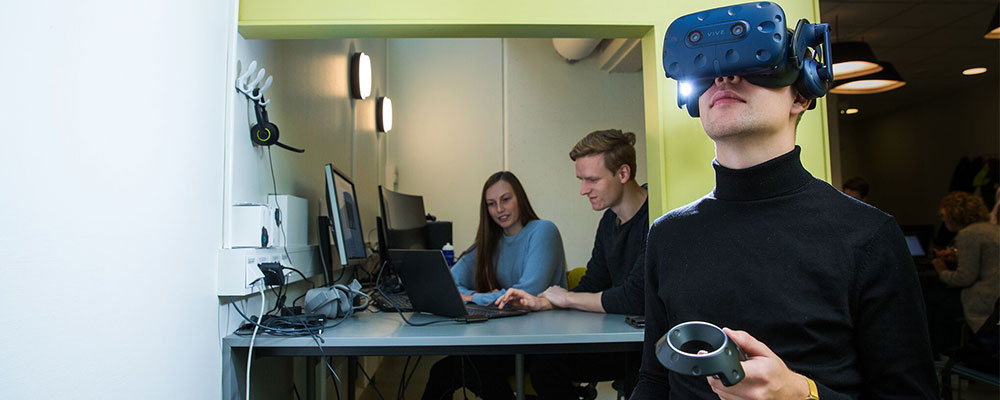 Student med VR-utstyr i forgrunnen, med to studenter som ser på en PC i bakgrunnen. Foto.