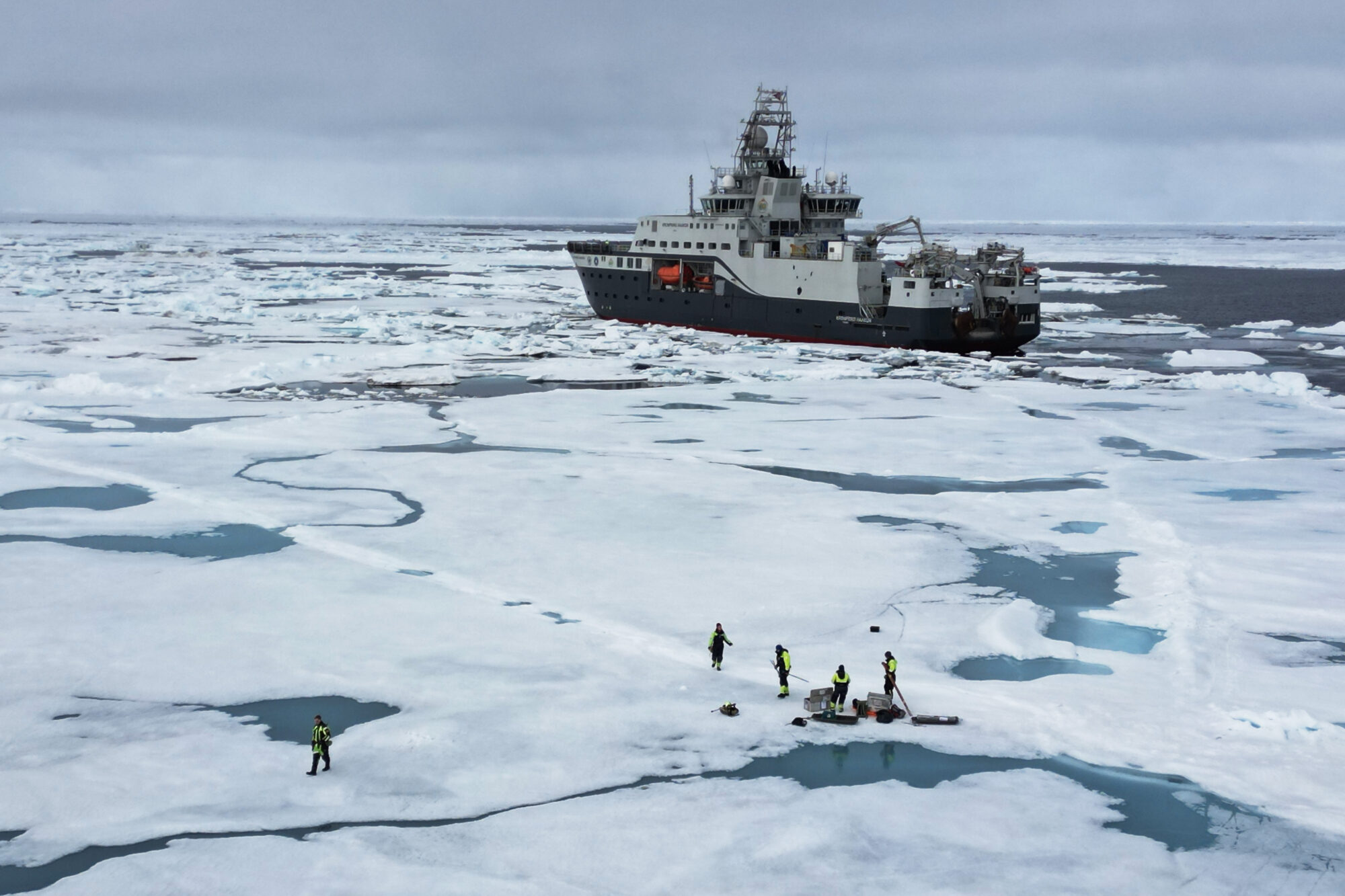Hav kledd med is. Fem personer står ute på isen med forskjellige instrumenter. Bak kan man se et skip