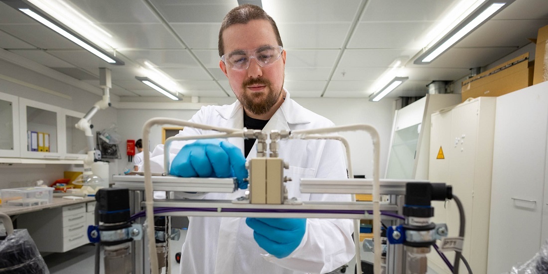 En mann står i et laboratorium. an har på seg vernebriller, blå hansker og hvit frakk. Hendene hans er på et apparat