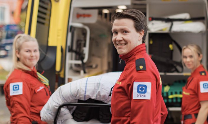 Tre personer i ambulanseuniform står bak en ambulanse og ser mot kamera