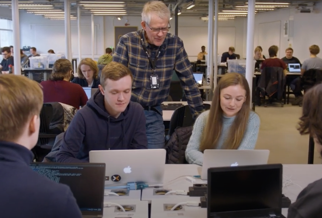 En lærer står bak to studenter som sitter med hver sin pc