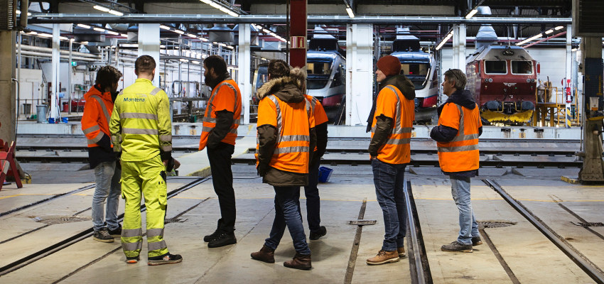 Sju mennesker i refleksvester oppholder seg i en toglokomotivhall. Foto