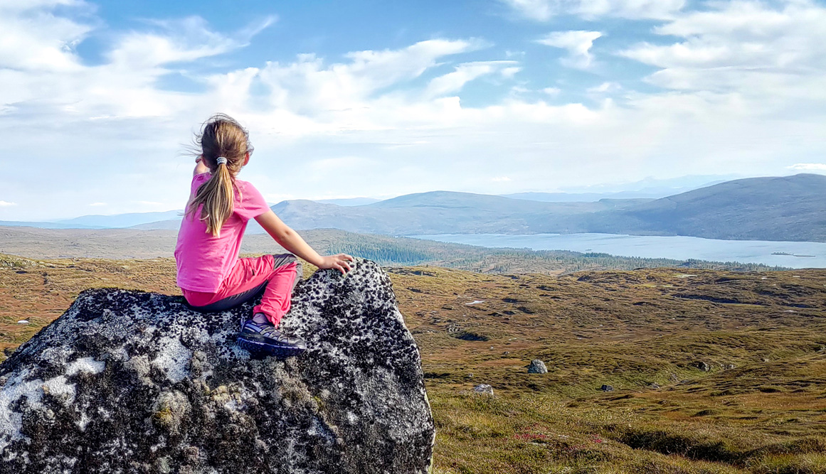 Ei liten jente som sitter på ei stein og peker fremover i et åpent landskap