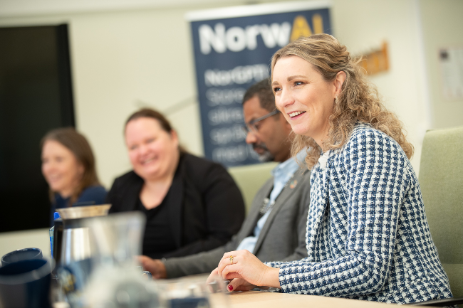 Fire personer sitter ved et bord under et møte med en ‘NorwAI’ plakat i bakgrunnen.