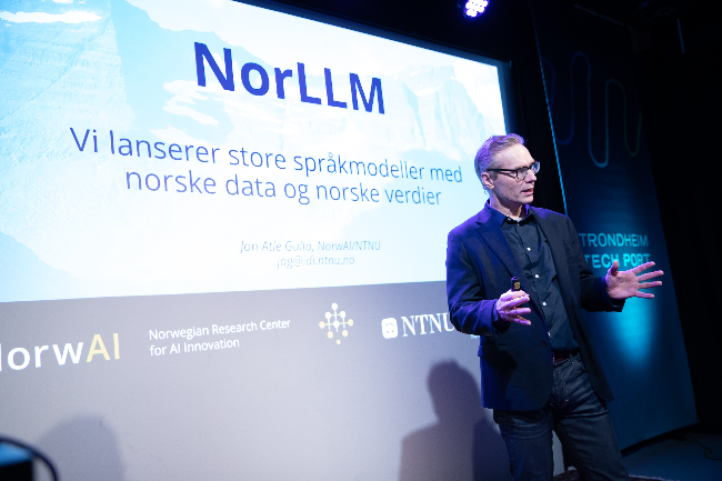 En presentasjon av NorLLM-prosjektet, som arbeider med store språkmodeller basert på norske data og verdier, på en konferanse i Trondheim. På skjermen vises informasjon om lanseringen av modellene og logoene til NorwAI og NTNU.