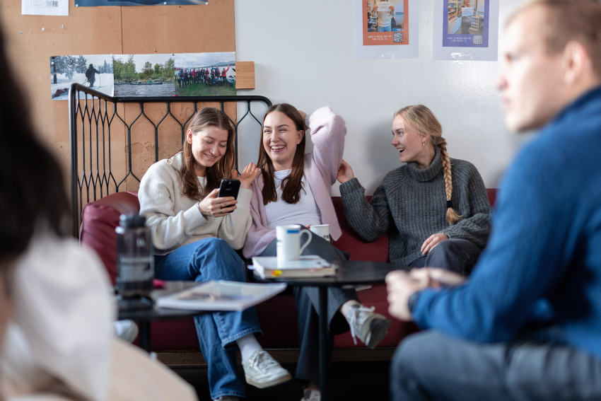 Tre kvinnelige studenter sitter sammen i en sofa, i et pauseareale for studenter, og ler av noe de ser på en mobil